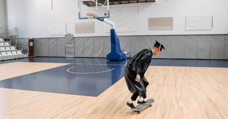 A graduate in a cap and gown skateboards on a basketball court indoors.